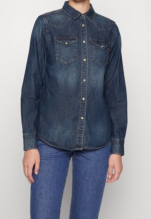 Person trägt ein langärmeliges dunkles Denim-Hemd mit zwei Brusttaschen und blaue Jeans, dargestellt vom Hals bis zur Hüfte.