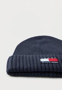 Temno modra pletena kapa z rebrasto teksturo in zavihanim robom. Na njej je pravokotna nalepka z logotipom v rdeči, beli in modri barvi.