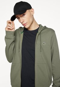 Olive žali zip-up džemperis su gobtuvu ir priekinėmis kišenėmis, dėvimas virš juodos marškinėlių, su mažuoju žaliu krokodilo logotipu. Juoda kepurė laikoma rankoje.