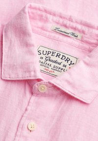 Chemise rose pâle à boutons avec des étiquettes indiquant "Laundered Wash" et "Superdry Graded 54 Coastal Supply Official Garment".