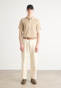 Polo beige, pantalon crème, ceinture en cuir marron, chaussures vert foncé et casquette noire. Le mannequin se tient dans un cadre neutre.