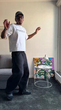 Fehér póló szöveggel, fekete melegítőnadrág, fekete szandál. A háttérben egy fehér lámpa és egy színes absztrakt festmény látható. Puha szőnyeg.