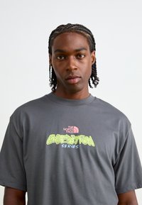 Grå t-shirt med korte ærmer, der har et pink The North Face-logo og lys grøn tekst "EXPEDITION SERIES" i fed skrift.