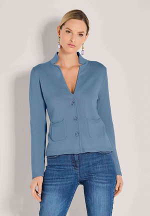 Frau in einem blauen, knöpfbaren Cardigan mit zwei Fronttaschen und blauen Jeans, stehend vor einem schlichten, hellen Hintergrund.
