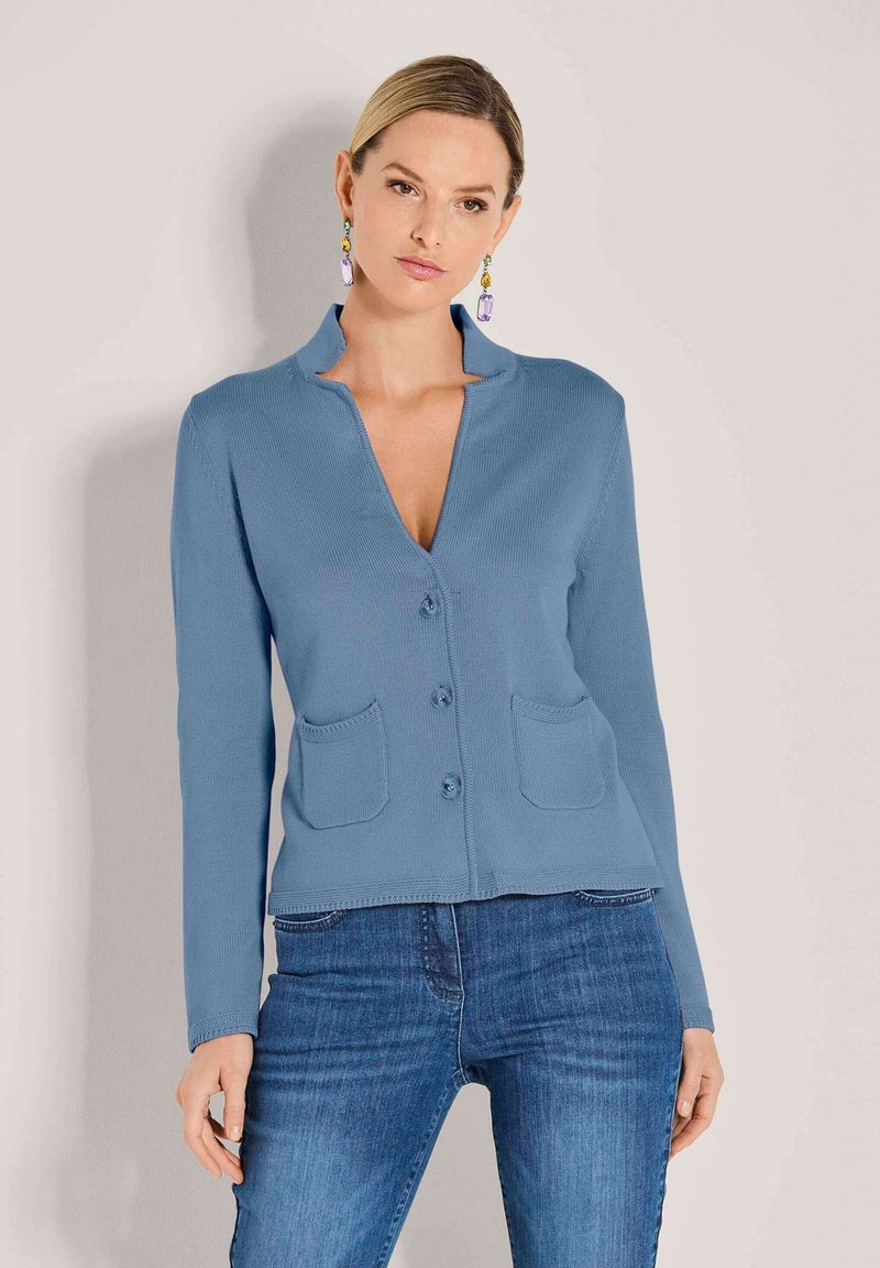 Vrouw in een blauwe knoopcardigan met twee voorzakken en blauwe jeans, staand tegen een effen lichte achtergrond.