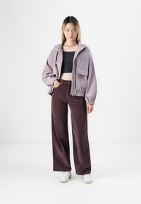Vrouw in een lavendel corduroy jasje, zwarte cropped top, donkerbruine wijde corduroy broek en witte sneakers, staand tegen een witte achtergrond.
