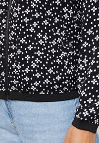 Veste bomber noire avec imprimé floral blanc, dotée d'une fermeture éclair, de poignets et d'un ourlet côtelés, associée à un jean bleu clair.