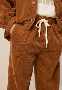 Ensemble en velours côtelé brun comprenant une veste à boutons et un pantalon assorti. Le pantalon possède une taille à cordon et une texture côtelée verticale.