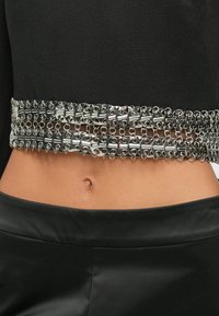 Μαύρη υφασμάτινη μπλούζα με ευρεία ασημένια αλυσίδα στη μέση, που διαθέτει διασυνδεδεμένα μεταλλικά κομμάτια και λεία υφή.