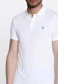 Moški nosi prilegajočo belo polo majico z modrim logotipom na levem prsnem delu, kar prikazuje njen preprost in čist dizajn.