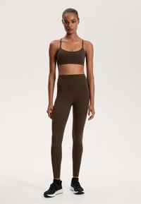 Hnědý sportovní crop top a legíny s vysokým pasem z elastického materiálu, doplněné černými teniskami. Hladká textura, minimalistický design.