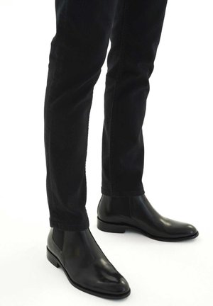 Bottes noires en cuir à cheville avec une texture lisse, un bout pointu, des panneaux élastiques sur les côtés et un talon bas, portées avec un jean foncé.
