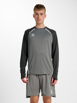 Junger Mann trägt ein graues und schwarzes langärmeliges Sportshirt und passende graue Shorts mit hummel-Logos auf Brust und Bein.