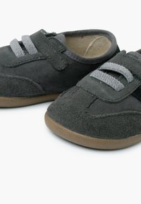 Scarpe per bambini di colore grigio scuro con lacci elastici grigio chiaro e cinturini in Velcro, texture in suede morbido e suole in gomma color beige su sfondo bianco.