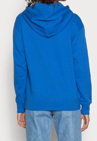 Felpa con cappuccio blu, dotata di tasca a marsupio frontale, polsini e orlo a costine. Tessuto morbido con una vestibilità rilassata, indossata sopra jeans di denim chiaro.