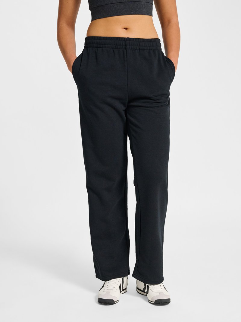Vrouw draagt zwarte joggingbroek met elastische taille en zakken, gecombineerd met witte en zwarte sneakers, cropped zwart topje, staand tegen een witte achtergrond.