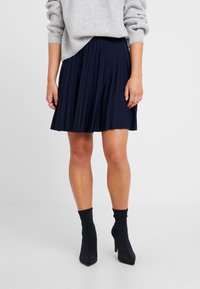 Marinblå plisserad kjol med en slät textur, tillsammans med tajta svarta ankelskor. Outfiten har en avslappnad grå tröja.