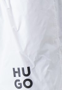 Bela tkanina z gladko, rahlo nagubano teksturo. Črn napis "HU GO" je izpostavljen. Dodatni oblikovni elementi niso vidni.