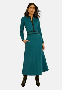 Rochie verde-albăstrui cu mâneci lungi, fermoar frontal, guler și talie negre, buzunare laterale și o siluetă fluidă. Textură netedă și lungime până la mijlocul gambei.