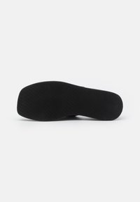 Semelle extérieure noire en caoutchouc d'une chaussure avec un motif de bande de roulement subtilement texturé, présentée à plat sur un fond blanc.