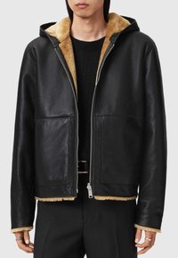 Schwarze Lederjacke mit Reißverschluss und Kapuze; verfügt über ein hellbraunes Lammfellfutter und -kanten, zwei Fronttaschen und einen lässigen Schnitt.