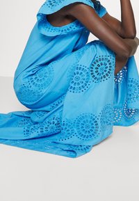 Robe en coton bleue ornée de broderies en dentelle aux motifs circulaires, avec une coupe décontractée et des boutons aux épaules, vue en position assise.