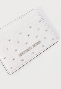 Άσπρο πορτοφόλι καρτών από δερμάτινη υφή με ασημένιες καρφίτσες. Διαθέτει λογότυπο "MICHAEL KORS" σε ασημί, τρεις θήκες για κάρτες και μαύρο περίγραμμα.