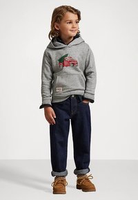 Vaikas, dėvintis pilką kapšelį su raudona sunkvežimio ir Kalėdų eglutės grafika, tamsias sukeltas džinsines kelnes ir rudus raišteliu užrišamus batus, stovi su atsipalaidavusiomis rankomis.