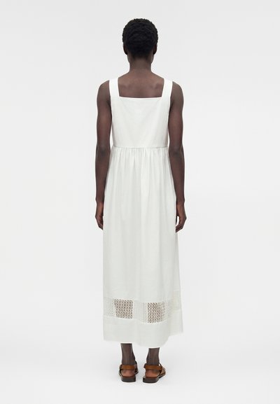 Άτομο με κοντά σγουρά μαλλιά που φοράει λευκό αμάνικο φόρεμα μήκους μίντι με δαντέλα κοντά στο κάτω μέρος, στέκεται γυρισμένο προς τα πίσω, φοράει καφέ σανδάλια.