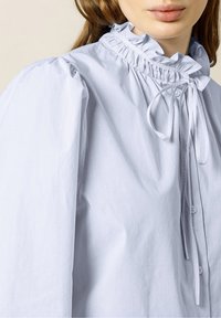 Camicia azzurro chiaro con colletto alto a volant, maniche a sbuffo e dettaglio con fiocco al collo; presenta sottili righe verticali.