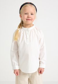 En ung flicka med blont flätat hår, iförd en vit knäppblus, ljusa byxor och ett mörkt blommigt hårband, står mot en enkel bakgrund.