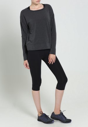 Persoon die een donkergrijze top met lange mouwen, zwarte cropped leggings en zwarte en paarse sportschoenen draagt, staat tegen een lichte achtergrond.