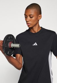 Svart idrottströja med en vit Adidas-logga, vertikala vita ränder på ärmarna, och en hand som håller en svart viktplatta med ett silverhandtag.