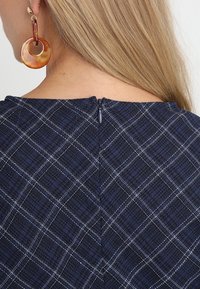 Mörkblå tyg topp med vit rutig mönster, utrustad med dragkedja bak och rund halsringning. Stora runda örhängen framhäver utseendet.