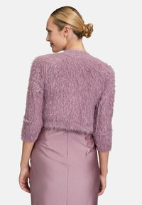 Cardigan court violet en peluche avec des manches trois-quarts, tissu texturé et coupe cropped, porté sur une jupe satinée coordonnée.