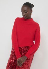 Pull rouge à col montant, à manches longues et à la texture douce, associé à une jupe rouge brillante ornée de sequins.