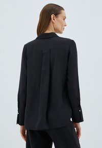 Camicia nera a maniche lunghe con colletto, con un dettaglio di cucitura sul retro e bottoni decorativi sui polsini, realizzata in tessuto liscio.
