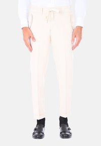 Pantaloni beige con vita regolabile, tasche laterali e vestibilità sartoriale. Indossati con scarpe nere senza lacci e calze nere.