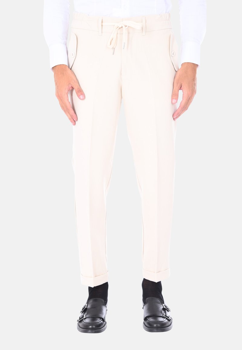 Pantaloni beige con vita regolabile, tasche laterali e vestibilità sartoriale. Indossati con scarpe nere senza lacci e calze nere.