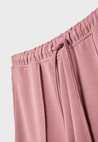 Svijetloroze trenirke s rastezljivim pojasom i podesivom vezicom, mekim teksturom tkanine i nabranim detaljima sa strane.