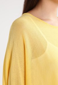 Gelbes, leichtes Strickoberteil mit lockerem, drapiertem Design und weitem Ausschnitt. Der Stoff hat eine leicht transparente Textur und eine glatte Oberfläche.