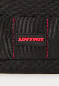 Zwarte stoffen tas met een gestructureerd oppervlak, een rode stiksel detail en een rechthoekige URTRN-logo patch in het rood op een zwarte achtergrond.