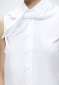 Gros plan d'une chemise blanche sans manches à boutons, avec une écharpe blanche nouée autour du cou et un texte brodé subtil sur la poitrine.