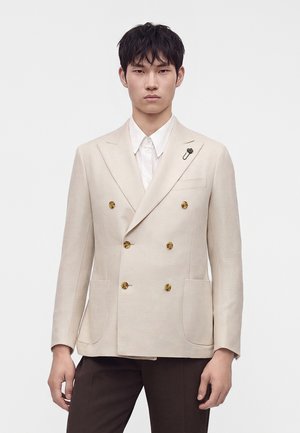 Mann trägt einen beigen zweireihigen Blazer mit goldenen Knöpfen, weißem Hemd und dunkelbrauner Hose und steht vor einem einfarbigen Hintergrund.