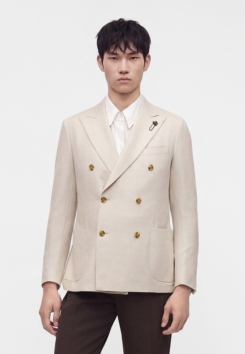 Homme portant un blazer beige croisé avec des boutons dorés, une chemise blanche et un pantalon marron foncé, debout devant un fond uni.
