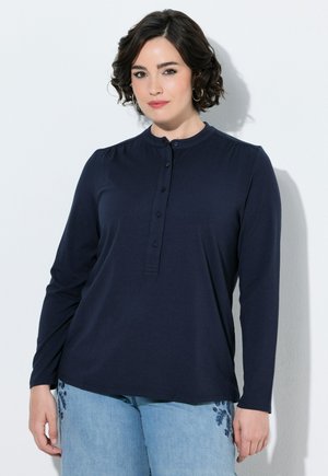 Donna con capelli corti, ricci e scuri, che indossa una camicia blu scuro a maniche lunghe con bottoni e jeans azzurri chiari con ricami floreali, in piedi davanti a uno sfondo bianco.