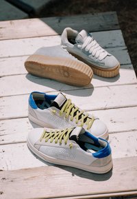Deux paires de baskets décontractées sur une surface en bois blanc, l'une avec des semelles épaisses en gomme, l'autre avec des accents bleus et des lacets jaunes.