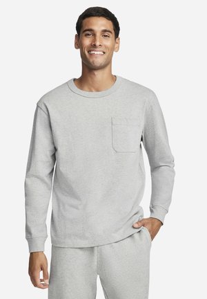 Man in light grey long-sleeve shirt with chest pocket and matching pants, smiling, with one hand in pocket against plain background.