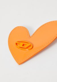 Hjärtfomad föremål i orange av glänsande material, med kontrasterande sömmar och en liten gul-orange logotyp i mitten.