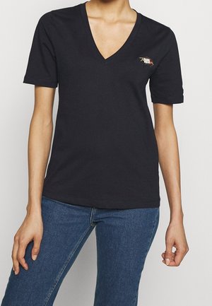 Mujer con camiseta negra de manga corta y cuello en V con pequeño diseño bordado en el pecho, combinada con vaqueros azules sobre fondo blanco.